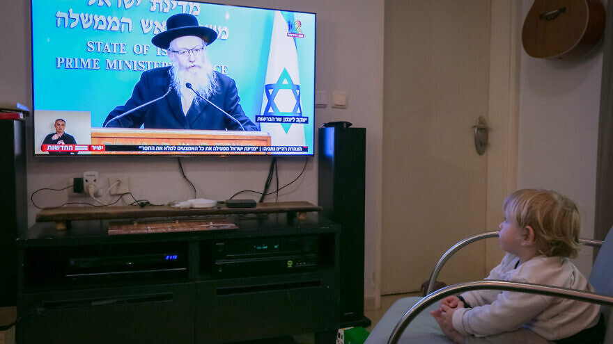 한 아이가 거실 의자에 앉아 TV를 보고 있습니다. TV 화면에는 이스라엘 공영방송 KAN의 메인 뉴스가 방송되고 있으며, 화면 왼쪽 하단에 수어 통역사가 수어 통역을 제공하는 모습이 함께 보입니다.