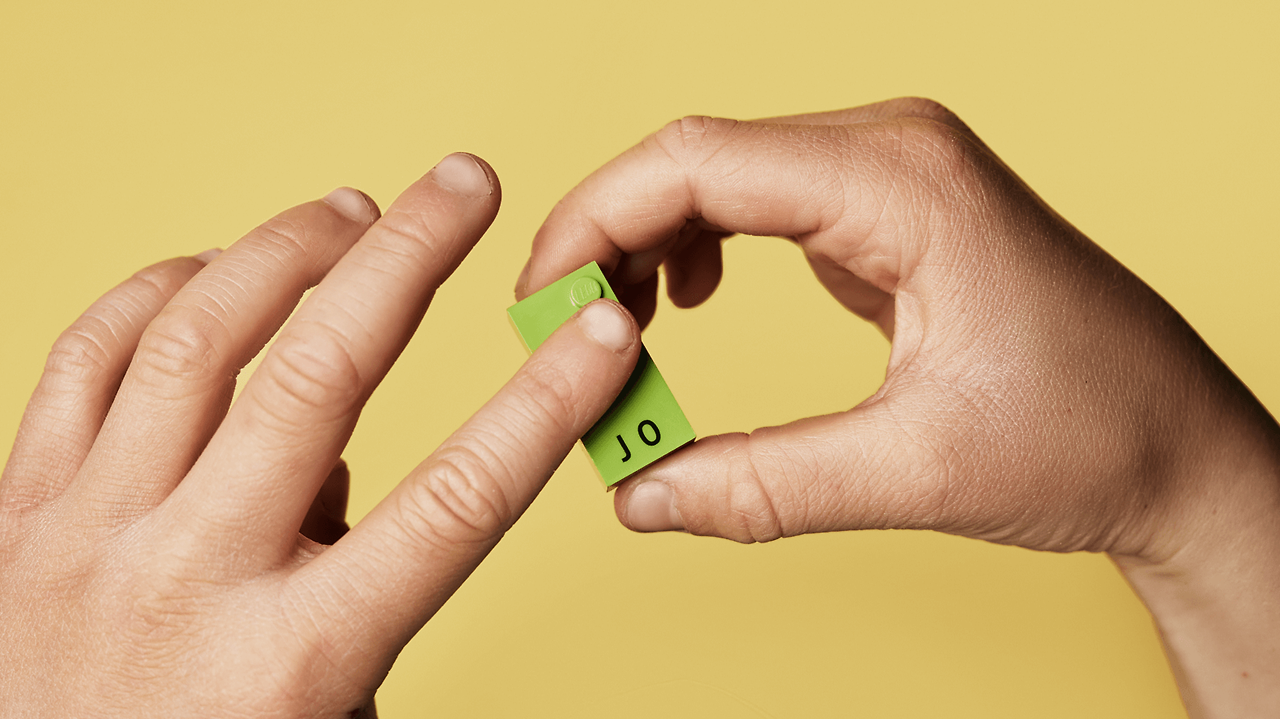 한 사람의 손이 'JO'라는 글자와 그에 해당하는 점자가 함께 인쇄된 녹색의 '레고 점자 블록' 하나를 들고 있는 클로즈업 사진입니다.